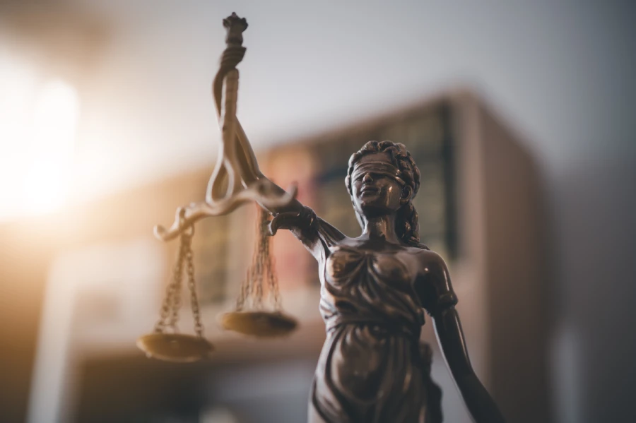 A bronze Lady Justice statue with a blindfold and balanced scales, highlighted by dramatic lighting against a dark, out-of-focus backdrop.