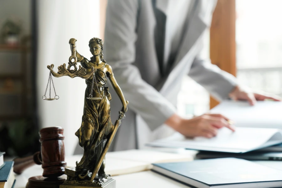 Bronze scales of justice in a sunlit office, with a lawyer in a gray suit handling papers in the background.