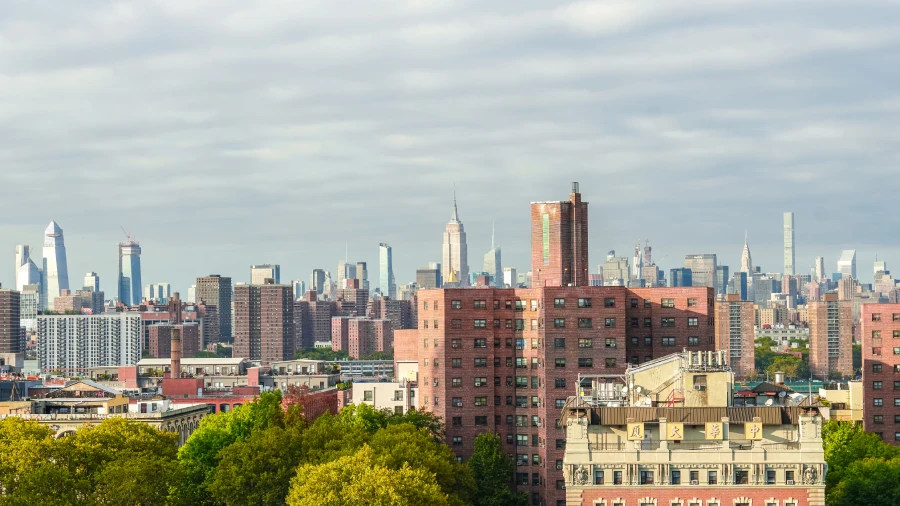 An aerial view of a city transition, showing red brick residential buildings and green trees in the foreground leading toward a dense skyline of modern glass skyscrapers under a soft, overcast sky.