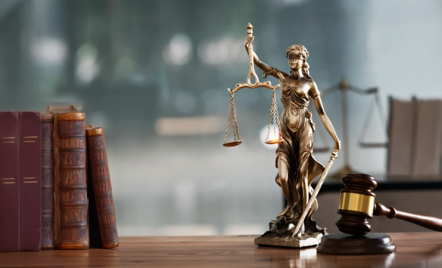 A bronze statue of the Scales of Justice stands balanced next to a wooden gavel with a gold band and a stack of leather-bound law books. The set is arranged on a dark wood table against a soft, blurred grey background.